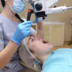 Young woman dentist treating root canals using microscope in the dental clinic. Man patient lying on dentist chair with open mouth. Medicine, dentistry and health care concept. Dental equipment